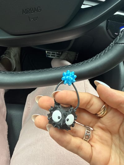 The scene is indoors, inside a car, likely in Santa Clara Ocoyucan, Mexico, as indicated by the broader context. A person is holding a hair tie with two decorative elements: one is a spiky black ball with large white eyes, reminiscent of a "soot sprite" or "dust bunny" character; the other is a spiky blue sphere. The person is wearing rings on two fingers: one a thick gold band and the other a silver band with Roman numerals. The car's steering wheel and airbag cover are visible in the background, as well as a portion of the pedals. The person is wearing a pale pink garment.