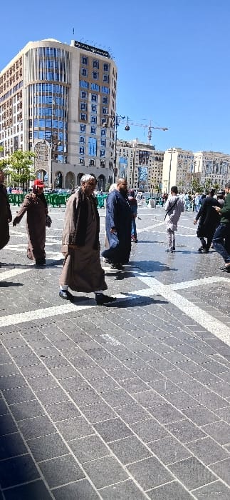The image displays a daytime view of a paved pedestrian plaza in Medina, Saudi Arabia, located in proximity to Al-Masjid an-Nabawi. The sky is clear and blue, and the scene is brightly illuminated by direct sunlight, evident from prominent shadows cast on the pavement. In the left background, a multi-story, modern building with a light-toned exterior and numerous blue-tinted, reflective windows stands tall; other contemporary structures and distant construction cranes are also visible. The plaza surface is composed of dark grey rectangular paving stones with lighter grout lines, forming a grid pattern. Approximately 10-15 individuals, predominantly adults, are observed walking across the plaza in various directions. Many are attired in traditional Islamic clothing, such as men's thobes and women's abayas. In the central foreground, a man with grey hair and a beard, wearing a light brown thobe and a dark scarf, walks towards the right. To his immediate left, another man in a dark brown thobe and a red cap proceeds leftwards. Green metal barriers are discernible in the mid-left background.