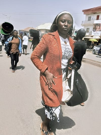 The image depicts a bustling outdoor street scene under bright daylight, characteristic of an urban or semi-urban environment. The specific location of Kungulu School, Zambia, is not identifiable within the visual context of the photograph; however, the setting appears to be a commercial thoroughfare in an African city.
In the foreground, a dark-skinned adult female stands facing the camera with a neutral, direct gaze. She wears a white headband, large circular hoop earrings, and a white t-shirt with visible dark text ("THIS GIRL IS NOT AFRAID TO SHINE"). Over the t-shirt, she wears a textured, orange-red blazer or long coat. A black and white patterned skirt or wrap extends below the coat, and she wears brown flip-flops. Her left hand rests on her hip, while her right hand holds a dark object, possibly a phone or wallet, near a white shoulder bag with a textured flap and metal clasp slung over her right side. A light-colored pendant hangs from a necklace.
Several individuals are visible in the background, walking away from or across the camera's perspective. To the left, a woman in a brown t-shirt and light blue jacket walks, carrying a dark shoulder bag. Further back, a person carries a large, dark green rolled item, and another carries a black bucket on their head. A male figure in a white t-shirt and dark shorts walks in the mid-ground. To the right and behind the foreground subject, a dark-skinned individual in a black shirt carries a large black bag or bundle.
The ground is a light grey paved surface, likely asphalt or concrete, with strong shadows indicating overhead sunlight. In the background, buildings are visible under a clear, pale blue sky. On the right, a two-story white building with orange-red trim features balconies and a large advertisement billboard. Other less distinct buildings appear to the left. Overhead power lines are visible. A light-colored market umbrella and distant vehicles are also present on the right, suggesting a busy commercial or market area.