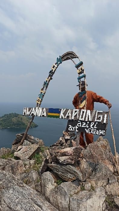 A single male individual stands on a rocky outcrop in Bwishyura, Rwanda, overlooking a large body of water and a distant, verdant island, beneath an overcast sky. The man is positioned behind a rustic wooden sign structure that includes a curved archway decorated with light blue and yellow elements. A prominent horizontal wooden sign reads "KWANIA KARONGI" in white lettering, featuring a painted Rwandan flag between the two words. Below this, a smaller black wooden sign with white text identifies "azizi art village." The man wears an orange-brown long-sleeved shirt and matching trousers, a dark head covering, and a multi-colored necklace. He holds a wooden staff in his right hand, resting it on the fragmented grey and brown rocks, while his left hand is placed on his hip. Sparse green vegetation grows between the rock formations. The body of water appears dark blue, and the distant island is heavily treed.