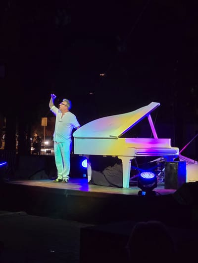 In this photograph, a man in a white outfit, with glasses and graying hair, is captured in mid-performance. He is positioned horizontally, as if flying or levitating, with his arms outstretched. He appears to be a performer on a stage, possibly a pianist, given the presence of a large, white, unconventional piano behind him.

The piano itself is a striking object, its sleek, modern design illuminated by vibrant stage lighting that casts colorful hues of purple, pink, and yellow across its surface and the surrounding area. Behind the piano, stage equipment is visible, including lighting fixtures, one of which emits a bright blue light.

The scene is set at night, with the darkness of the background emphasizing the illuminated stage and performer. Hints of an outdoor setting can be seen in the background, with faint lights and structures suggesting it might be an open-air event. The lighting creates a dramatic and theatrical atmosphere, hinting at a captivating performance. The overall mood seems energetic and perhaps a bit surreal due to the man's unusual posture and the artistic presentation of the piano. There is no visible text in the image.