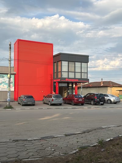 This image captures a street scene in Saedinenie, Bulgaria, featuring a modern commercial development adjacent to an older residential structure. In the foreground, a gray asphalt road runs horizontally, bordered by a curb and a sidewalk paved with hexagonal gray concrete blocks, interspersed with sparse weeds.

The central focus is a multi-part commercial building. To the left, a tall, plain, rectangular structure is clad entirely in bright red corrugated material. Immediately to its right is a two-story building with a dark gray facade and prominent windows on the upper floor. This building features a red overhang with the Cyrillic text "Фаворит" (Favorit) in white script, indicating a store or supermarket. Below "Фаворит," another sign reads "Супермаркет" (Supermarket). A red support column stands at the entrance.

Parked in front of the "Фаворит" building are several cars. From left to right, visible are: a dark gray Opel Astra hatchback with license plate "CT 1146 PT," a silver station wagon with license plate "PB 5304 HB," a red Chevrolet Lanos/Aveo sedan with license plate "PB 6001 BA," a black Toyota RAV4 SUV with a rear-mounted spare tire and license plate "PB 3117 TE," and a silver Cadillac sedan.

To the far left, partially visible, is another building with a green billboard featuring white text in Bulgarian, including "ДОБРИЯТ ИЗБОР ЗА ЦЯЛОТО СЕМЕЙСТВО" (The good choice for the whole family) and imagery of packaged goods. A gray utility pole with various attached wires stands prominently in front of this billboard. Overhead, numerous power lines stretch across the scene.

A single person, a male in an orange t-shirt, is visible standing near the entrance of the "Супермаркет."

Behind the commercial buildings, an older, single-story house with a weathered tiled roof and a brick chimney is visible. The sky above is mostly overcast with scattered white and gray cumulus clouds, suggesting either late morning or early afternoon with soft, diffused sunlight. The overall impression is a mix of contemporary commercial development and older infrastructure in a suburban or semi-urban setting.