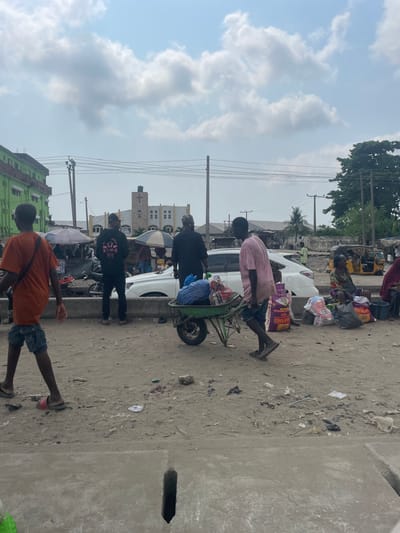 The image presents a busy outdoor market scene in Ojo, Nigeria, under a partly cloudy sky. The immediate foreground consists of uneven, light-brown dirt ground, scattered with debris, and a concrete slab featuring a dark, rectangular drain opening.
A young male in an orange t-shirt and blue shorts, wearing a dark flip-flop on his right foot and a reddish-pink one on his left, walks towards the left of the frame. Behind him, a male in a black hoodie with a white graphic print on the back stands facing away from the camera. Adjacent, a white SUV is parked, and a male in a dark garment and cap stands nearby. A male in a light pink t-shirt pushes a green wheelbarrow loaded with blue and white sacks towards the right.
On the right side of the frame, several individuals, predominantly women, are seated on the ground amidst numerous large sacks, some labeled "Quality Rice" and "PEAS," indicating market goods. Multiple yellow motorized tricycles (Keke Napep) are visible further back on the right.
In the background, a light-colored church building with a cross on its central steeple is prominently visible. To the far left, a multi-story building with a bright green facade is present. Utility poles with multiple overhead wires span the upper midground. The environment suggests an active urban commercial area.