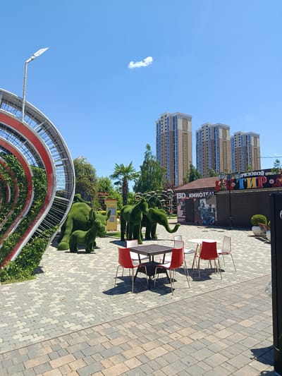 This is an outdoor photograph taken on a clear, sunny day. The scene depicts a paved plaza with several elements of decor and entertainment. In the foreground, there are two tables with chairs, some of which are red and white. Behind the tables, there are topiaries shaped like animals, including a large rhino with a smaller one, and an elephant. To the left, a large, curved metal structure covered in greenery is visible, resembling a tunnel or archway.

In the background, several tall residential buildings with blue windows and ornate designs on their facades stand against the bright blue sky. A smaller building with a sign that reads "3D КИНОТЕАТР" (3D CINEMA) and another sign with Cyrillic text, "РИЗОВОЙ ТИР" (Rizovoy Tir, likely a shooting gallery or entertainment center), suggests a recreational area. Palm trees and other greenery are scattered throughout the scene, indicating a warm climate. The sun is high in the sky, casting sharp shadows, suggesting it is midday. The overall atmosphere is one of a public space designed for leisure and entertainment.