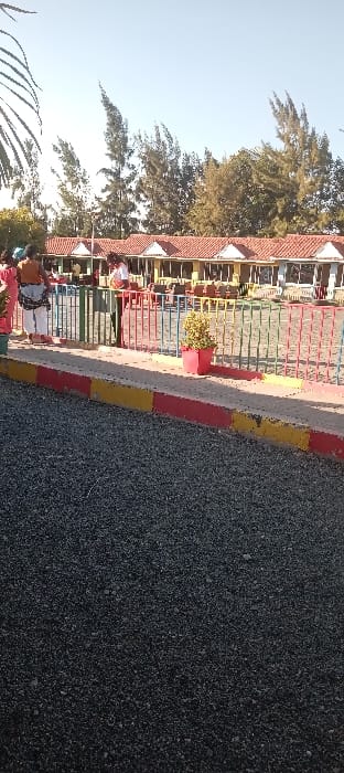 The image shows a view of a paved area in front of a row of buildings, likely a resort or hotel, in Athi River, Kenya. The buildings are yellow and white with red tile roofs. There is a colorful metal fence in front of them, with a small green bush and red flower pot. Three people are walking away from the camera towards the fence, and a fourth person is walking in the opposite direction. The ground is covered in small gravel, and there is a paved sidewalk with yellow and red markings. The trees in the background are green and lush. The sky is clear and blue.