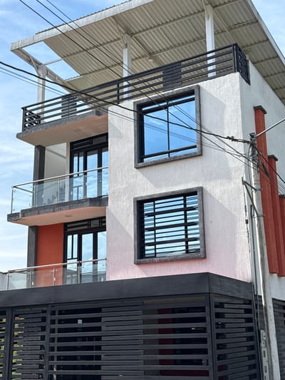 The image depicts a multi-story building in Pitalito, Colombia, viewed from a low angle. The structure features white textured stucco walls, with dark gray/black trim around windows and prominent dark gray/black structural pillars. On the lower two visible levels, there are balconies with clear glass and silver metal railings, alongside dark-framed glass doors. The right side of the building incorporates vertical reddish-orange textured panels.

The third level from the bottom features two rectangular windows; the upper one shows a reflection of the sky and a cross-pattern, while the lower one has horizontal blinds visible behind the glass. Above the third level, a balcony with a dark gray/black horizontal slatted metal railing is present. The building is topped with an open-air roof structure composed of a corrugated metal sheet canopy supported by white pillars.

At ground level, a large dark gray/black horizontal slatted metal gate spans the width of the building, providing access. Utility wires, black in color, traverse the upper portion of the frame and are attached to a utility pole on the far right, which also supports some of the reddish-orange panels. The sky is partially cloudy and bright. No individuals or active interactions are visible.