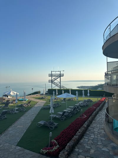 The image depicts an outdoor area with a view of the sea, likely from a resort or hotel in Topola, Bulgaria. In the foreground, there is a paved pathway leading past rows of empty lounge chairs and closed umbrellas, all set on a manicured lawn. A long, vibrant red flowerbed runs along the right side of the pathway. To the left, the lawn slopes down towards the sea, with more lounge chairs and umbrellas visible.
In the mid-ground, a black metal tower with multiple levels and staircases stands against the backdrop of the calm, blue sea and a hazy coastline. The sea stretches to the horizon, with a clear, light blue sky above. On the right side of the image, a curved building with balconies and glass railings is partially visible. The overall impression is one of a serene and well-maintained outdoor space designed for relaxation and enjoying the coastal scenery. There are no people visible in the image.