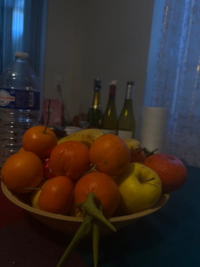 The image displays an indoor still life scene, reportedly located in Arthès, France. The foreground is dominated by a light-colored, possibly woven, bowl filled with various fruits. Prominently featured are multiple oranges or tangerines, some retaining small green leafy stems. Also present are at least one yellow apple, several yellow bananas, and one dark red, textured fruit, possibly a pomegranate, situated on the right side of the bowl.
To the left of the fruit bowl stands a partially full, clear 1.5-liter plastic bottle of "CRISTALINE" mineral water, identifiable by its blue and white label. The objects rest on a surface covered by a multi-colored fabric, exhibiting sections of dark red, purple, and dark teal.
In the mid-ground, behind the fruit bowl, three bottles are visible: a dark-colored bottle with a gold-foil top, likely sparkling wine; a dark green bottle with a red-foil top, likely red wine; and a lighter green bottle with a silver or white-foil top, likely white wine. A clear, empty stemmed glass is positioned in front of these bottles. Further to the right, a white cylindrical object, possibly a cup or vase, is present. A blurry pink rectangular object is also visible in the background, behind the water bottle.
The background consists of a light-colored wall, featuring an electrical outlet on the left. The scene is dimly lit, with a distinct vertical column of blue light visible on both the far left and right, suggesting an external light source filtering through a sheer, light-colored curtain on the right. No individuals are depicted in the image.