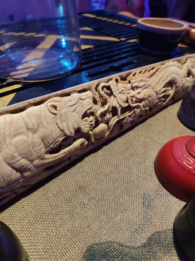 The scene is set in Mas, Indonesia. An elongated, pale-toned relief carving, likely wood or bone, occupies the central foreground, resting horizontally on a textured, light brown textile surface. The carving depicts a roaring tiger on the left, facing right, in dynamic interaction with a detailed dragon on the right, facing left. The tiger exhibits carved fur textures and an open mouth with visible teeth. The dragon features scales and sinuous lines. In the upper left background, a clear glass vessel exhibiting blue light reflections sits on a dark, grooved tray. To the upper right, a small, two-toned ceramic cup is partially visible on the tray. In the lower right foreground, a partial view of a red, hemispherical object and a dark, cylindrical container are present. Ambient lighting is subdued, with prominent blue illumination reflecting off the glass vessel. No individuals are depicted.