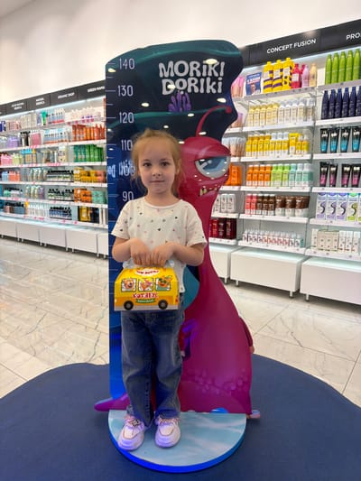 A young girl stands on a blue and white platform shaped like a circle, positioned next to a height chart with a cartoon monster figure. The chart measures height in centimeters from 90 to 140. She is wearing a white t-shirt with small heart patterns, blue jeans, and white sneakers. The girl is holding a yellow box with cartoon cats printed on it, with the text "ТРИ КОТО КОТЯБУС", which translates to "Three Cats Kotyabus", and "ЕДЕМ В ДУБАЙ", meaning "We are going to Dubai".
Behind her, shelves stocked with numerous hair products are visible. The shelves are labeled with brand names such as "CONCEPT FUSION", "PRO", "GREEN MAMA", "ORGANIC SHOP", "NATURA SIBERICA", and "ICE BY NATURA SIBERICA".
The scene is brightly lit, and the floor is patterned with light-colored tiles. The location is a retail store in Novoaleksandrovka, Russia, likely a cosmetics or personal care store.