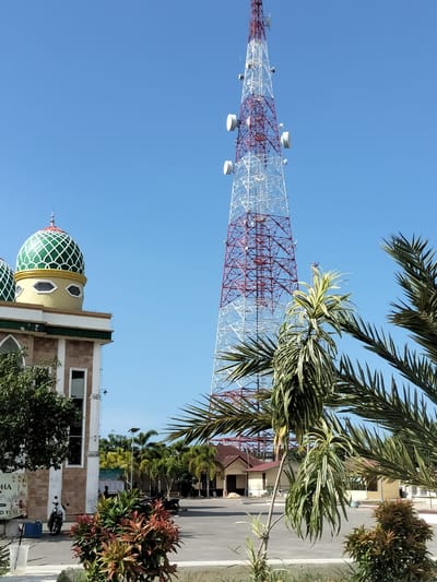The image shows an outdoor scene in Gampongjawa, Indonesia, under a clear blue sky. A tall, lattice-structured telecommunications tower dominates the background, colored red and white, with cylindrical microwave dishes attached at various heights. To the left stands a portion of a building, likely a mosque, with a green, patterned dome visible. In the foreground, various plants and trees are present, partially obscuring a view of a paved area that contains parked motorcycles and what seems to be a small, one-story building. A few people are visible in the middle ground, though their actions are unclear.