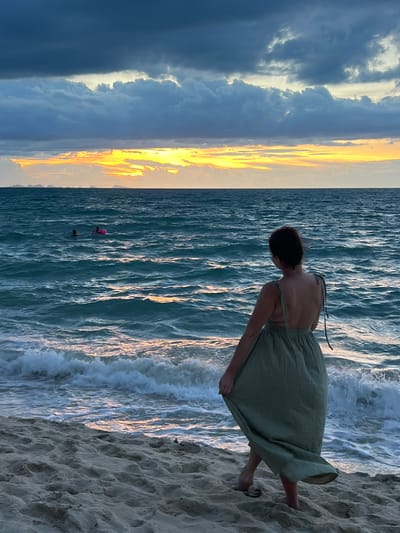 A woman stands on a sandy beach in Baan Tai, Thailand, facing the ocean at sunset. She wears a light green, flowing dress that trails behind her as she lifts the hem with her left hand. Her back is to the viewer, and her hair is dark brown and pulled back. In the distance, two figures can be seen swimming in the choppy, teal-colored water. One figure appears to be wearing a pink inflatable ring. The sky above is filled with dramatic clouds, with a sliver of bright orange and yellow sunset visible on the horizon, peeking through a break in the dark grey clouds. The waves are crashing onto the shore, creating white foam. The sand is a light beige color, with subtle footprints visible. Several small islands or landmasses are faintly visible in the far distance.