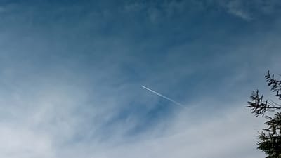 The image shows a light blue sky with scattered clouds. A jet contrail stretches diagonally across the center of the frame. A tree branch with leaves is visible in the lower right corner. The location is Ndeiya, Kenya.