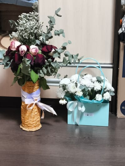 Two floral arrangements are displayed on a dark wooden floor in front of a beige-colored wall. To the left, a bouquet of red and pink roses with eucalyptus leaves is arranged in a tall, textured, woven vase. The vase is adorned with two ribbons, one lavender and one white, tied in a bow at the front. To the right, a light blue gift bag holds a dense bouquet of white chrysanthemums, also with eucalyptus leaves. The gift bag has two light blue handles and a white ribbon tied in a bow on the front. A small label with the word "Avocado" is visible on the front of the gift bag. The background wall appears to be a door or cabinet with vertical lines. There is no discernible information to indicate the specific location within the city of Pavlodar, Kazakhstan.