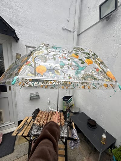 A low-angle shot depicts an outdoor cooking scene in Gorleston-on-Sea, United Kingdom. An individual's arm, clad in a brown jacket, reaches towards a barbecue grill loaded with several skewers of meat. The grill sits on a wooden stand, and a fork and a pair of tongs rest on a lower rack. Behind the grill, a white wall is visible. To the left, a clear umbrella with a floral and hummingbird pattern hangs from a clothesline. A portion of a table with items on it is visible in the upper-right corner. A window and door are at the bottom. The scene is situated in Gorleston-on-Sea, United Kingdom.