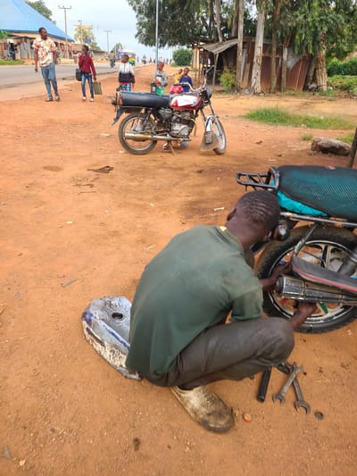 In Vom, Nigeria, a man is crouched on reddish-brown dirt, working on the rear wheel of a motorcycle. He wears a dark green shirt and dark pants. Tools, including wrenches, lie on the ground near him. A detached motorcycle fuel tank, a blue and silver metal object, rests on the dirt to his left. Behind him, another motorcycle, predominantly red with black accents, is parked on the dirt. In the background, a street runs horizontally, with people walking on the left side. A building with a blue roof and other structures line the street, amidst trees and vegetation. The sky is overcast.