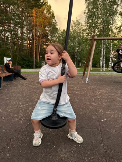 An outdoor shot captures a young girl on a swing set at a playground, likely in Surgut, Russia. The girl, with light brown hair and an open mouth, is seated on a dark, circular swing. She wears a white t-shirt with a subtle design and light blue denim shorts adorned with white butterfly patterns. Her feet are clad in white and pink sneakers with pink socks peeking out. In the background, a young person in a black t-shirt is visible on another swing. To the left, a man in a black t-shirt and pants sits on a wooden bench, looking at his phone. The playground surface appears to be dark brown with scattered debris. The environment is characterized by a backdrop of tall, slender trees with lush green foliage, and a pale, hazy sky suggesting either dawn or dusk.