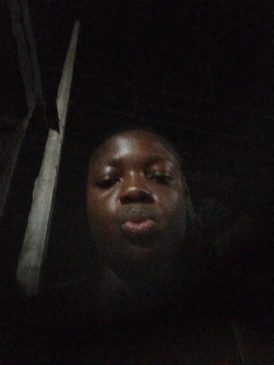 A close-up selfie of a person with dark skin is the main subject of the image. The person is looking directly at the camera with a neutral expression. The background is very dark, making it difficult to discern any specific details. A blurry object resembling a pole or piece of wood is visible on the left side of the frame. It is difficult to ascertain further details or location in Mbribit, Nigeria due to the poor lighting and limited visibility.