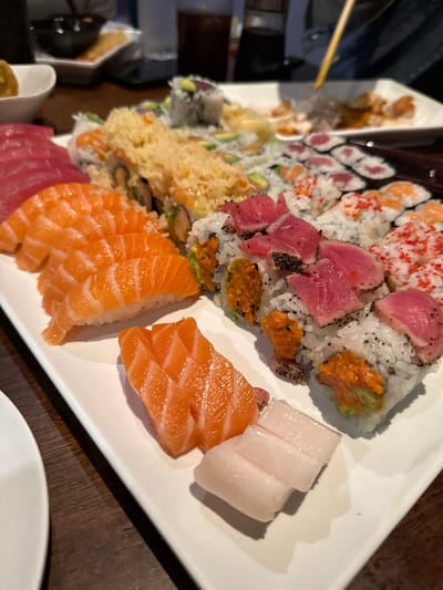 The image displays a large, rectangular white platter laden with a comprehensive assortment of sushi and sashimi, positioned on a dark wooden table in an indoor restaurant setting. On the left side of the platter, multiple slices of red tuna sashimi are visible, followed by several pieces of salmon nigiri, each consisting of a salmon slice atop a bed of rice. Below these are additional cuts of salmon sashimi and two pieces of white fish sashimi. The central and right sections of the platter are filled with various sushi rolls, including some topped with golden-brown tempura flakes, others with seared, pepper-crusted tuna, and rolls featuring vibrant red roe. Additional rolls contain ingredients like avocado. In the blurred background, several white dishes, dark beverages in glasses, and a human hand holding chopsticks, actively engaging with food on another plate, are discernible. The environment features subdued lighting. The specific restaurant in West Windsor, United States, is not identifiable from the visual content.