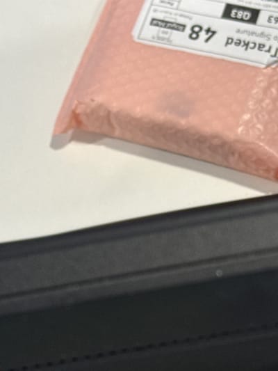 The image depicts a pink padded envelope with a shipping label. The label indicates "Tracked 48" and the code "Q83." Part of a black, textured object is visible on the right. The surface underneath is white. There's no indication of location in Hull, United Kingdom.