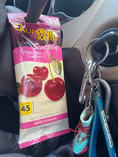 The image shows a close-up of a car's interior, specifically the steering column area. A pink and white air freshener labeled "California Scents Scent Bloc Coronado Cherry" is hanging from the steering column. Next to the air freshener, a set of keys hangs from the ignition. Attached to the keys are several keychains, including a small turquoise sneaker with red laces and a blue keychain with yellow lettering. The overall lighting suggests it is daytime. The specific location of Balch Springs, United States of America, is not discernible from the image itself.