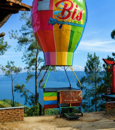 The image shows a colorful hot air balloon replica situated at a scenic overlook in Parik Sabungan, Indonesia. The balloon is predominantly red, yellow, green, blue, and purple, with the word "Bis" emblazoned in red script across the top. Beneath it, the text "Bukit Indah Simarjarunjung" is written in blue. A small, multi-colored basket hangs from the balloon, featuring red, yellow, blue, and green stripes. A sign attached to the basket reads "Sepeda Motor" with an arrow pointing left. The hot air balloon is positioned on a dirt path overlooking a vast blue lake and rolling green hills under a clear blue sky with scattered clouds. In the foreground, to the left, is a short brick wall. To the right, a red torii-style gate and a small structure are visible. Pine trees surround the overlook. There are no people or animals in the image.
