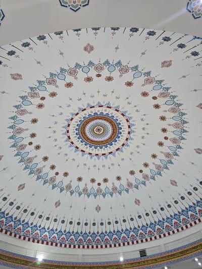 An indoor photograph taken from below shows the intricately decorated dome of a building. The dome is painted white with elaborate geometric and floral patterns in blue, red, gold, and green. The central part of the dome features a circular mandala-like design with calligraphy. Surrounding this central motif are concentric rings of smaller star-shaped and abstract decorative elements. Towards the bottom of the image, a decorative frieze with Arabic calligraphy runs along the edge of the dome. The lighting suggests a bright, possibly daytime interior. The context provided indicates the location is Paya Tringgadeng, Indonesia. There are no people or visible actions depicted.