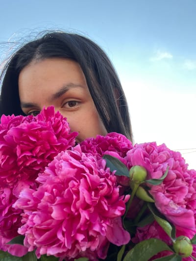An extreme close-up, low-angle shot captures a young woman partially obscured by a vibrant bouquet of bright pink peonies, held up to her face. Her dark hair cascades down, framing a portion of her face, with her left eye and eyebrow clearly visible. The background consists of a bright, pale blue sky with a few wispy white clouds. The lighting suggests an outdoor setting during the daytime, possibly in Oktyabrsky, Russia, given the context. The focus is sharp on the flowers and the woman's visible features, creating a sense of intimacy and vibrancy.