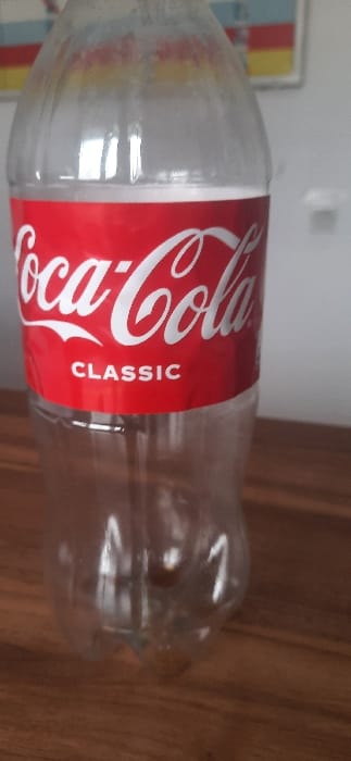 An empty, clear plastic Coca-Cola "CLASSIC" bottle stands upright on a brown wooden surface. The bottle's red and white label is clearly visible. Interior walls of the bottle show signs of residual moisture or film. The background consists of a light-colored wall, with a partially visible, blurred framed object displaying horizontal stripes of red, white, and yellow in the upper left. The scene is located in the city of Qorovulbozor, Uzbekistan. No individuals or specific actions are depicted.