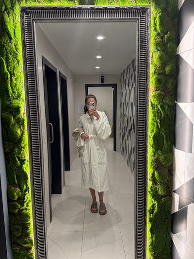 A person, wearing a white robe and sandals, stands in a modern, brightly lit room, taking a selfie in a large mirror. The room features a distinctive design with a ceiling adorned with a black and white geometric pattern and a floor covered in large white tiles with gray veining. The mirror itself is ornately framed with a black and white patterned border and is surrounded by a vibrant display of green moss. In the reflection, the person is holding a phone and appears relaxed. The space seems to be a spa or wellness area, possibly a changing room, given the robe and the clean, minimalist aesthetic. The overall impression is one of modern luxury and natural integration.