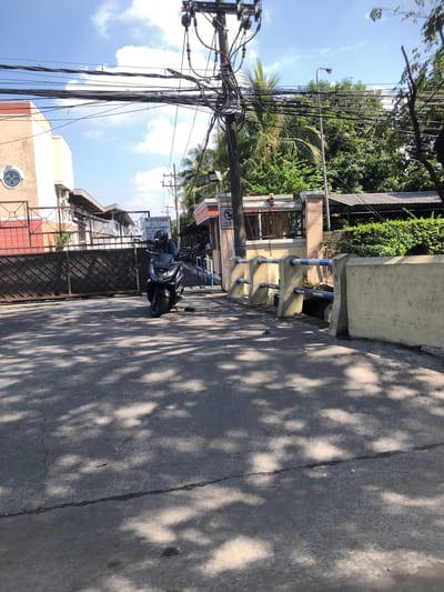 The image depicts a street scene in Banjarkemantren, Indonesia. A person wearing a helmet is riding a dark-colored motorcycle. The motorcycle has license plate "AD 3478 ZE". The rider is positioned near a large gate. In the background, there is a building with a cream-colored facade and a brown fence. Electrical wires and utility poles are visible overhead, with palm trees and other lush greenery surrounding the area. A sign with text in Indonesian is posted on a pillar. The road surface is asphalt, with shadows from trees cast across it. The sky is blue with scattered clouds.