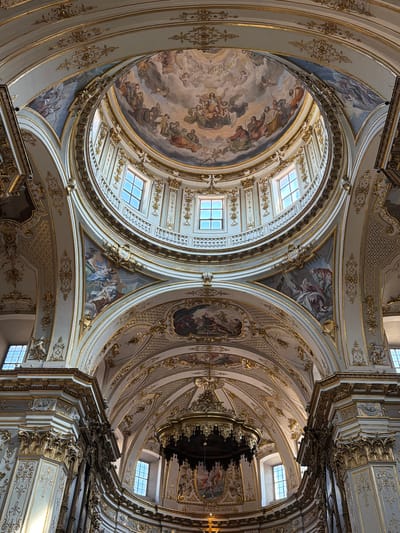 This media file depicts the ornate interior of a cathedral dome, viewed from below. The scene is dominated by elaborate Baroque architecture, characterized by its rich gold detailing, white plasterwork, and intricate frescos adorning the curved surfaces. Two large domed sections are visible, each featuring detailed ceiling paintings depicting biblical or mythological scenes with numerous figures. Skylights are integrated into the domes, allowing natural light to illuminate the vast interior space. The ceiling is further embellished with decorative moldings, gold leaf accents, and sculpted elements, creating a sense of opulence and grandeur. Suspended from the ceiling, a dark, intricate object, possibly a chandelier or part of the architectural structure, casts shadows. The overall impression is one of awe-inspiring artistry and religious devotion. The location is identified as Mozzo, Italy. There is no discernible text within the image.