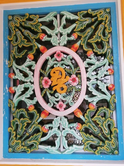 The image shows a close-up of an ornately carved window grill. The grill is made of wood and painted with vibrant colors, predominantly green, yellow, light blue, and pink. It features intricate floral and foliate motifs, with some elements resembling buds or small fruits. In the center of the grill, there is a pink oval frame containing a gold-colored Chinese character. The window is set within a blue frame, which is itself bordered by a yellow frame. The background seen through the grill appears to be a window pane reflecting trees and some architectural elements, suggesting an outdoor setting. The style of the carving and the vibrant colors are characteristic of traditional Southeast Asian architecture, possibly found in a temple or a heritage building in the city of Xuân Thành, Vietnam.
