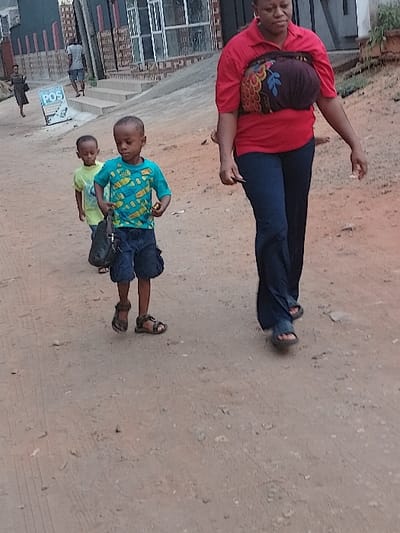 The image depicts an outdoor scene in Orie Uratta, Nigeria, on an unpaved, reddish-brown dirt path. In the foreground, an adult female with dark complexion walks towards the left, wearing a red short-sleeved collared shirt, dark blue trousers, and dark sandals. A dark, patterned fabric with colorful feather and leaf motifs is wrapped around her torso. She holds a small, dark object in her left hand.
Behind her, two young boys follow. The boy closer to the foreground, approximately 5-7 years old, wears a turquoise t-shirt with yellow fish patterns, dark blue shorts, and brown sandals. He carries a black bag in his left hand and a small, light-colored object in his right. The second, younger boy, partially obscured, wears a light green t-shirt and dark shorts.
In the mid-ground, an adult male in a light grey t-shirt and dark shorts walks away from the viewer. Further in the background, another adult female in a dark dress is also walking.
The background consists of several buildings, some with windows, and a set of concrete steps leading up. A white rectangular sign displaying "POS" in blue text is visible on the left side of the path. The ground is uneven, dotted with small stones, and exhibits faint tire tracks. Minimal green vegetation is present on an elevated area to the upper right. All visible individuals are in motion, moving generally from right to left across the frame.