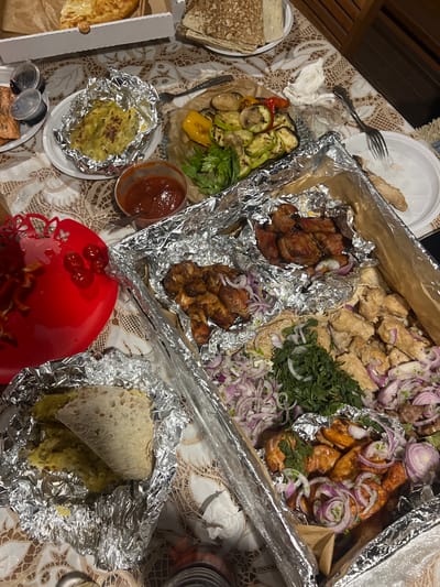 The image captures a top-down view of an abundant meal laid out on a table, covered by a patterned, light-colored tablecloth, likely indoors. The scene suggests a casual dining event or a large takeout order.

The central subjects are a variety of cooked dishes, predominantly meats and vegetables, presented in large aluminum foil-lined containers and on plates. Several sections of grilled meats are visible, including what appears to be shashlik (skewered meat pieces) and grilled chicken parts, all generously garnished with thinly sliced red onions and fresh herbs like cilantro or parsley.

Alongside the meats, there is a plate featuring an assortment of grilled vegetables, including zucchini, bell peppers (yellow and red), and mushrooms. Other food items include flatbread (likely lavash), some stacked on a white plate, and one portion used to wrap a mashed potato-like dish, also encased in foil. Another serving of this potato dish is presented on a small white plate, wrapped in foil. Various condiments are present, including a red, possibly tomato-based sauce in a clear plastic cup and smaller plastic containers with a darker sauce. A decorative red plastic dish holds a few cherry tomatoes and some chopped red peppers. Part of a pizza box or similar food packaging is visible on the left edge.

The activity taking place is a meal, likely shared by multiple people, given the quantity and variety of food. Notable details include the casual presentation using disposable white plastic plates, a metal fork, and crumpled napkins. The ambient lighting suggests it is evening or nighttime. No people are visible in the frame, and there is no visible text. The style of food, particularly the grilled meats and lavash, aligns with culinary traditions found in the given location context of Zima Yuzhnaya, Russia.