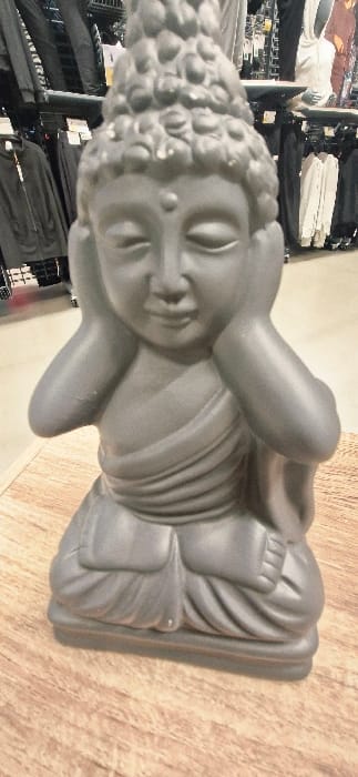 A dark grey Buddha statue, seated in a lotus-like position with hands covering its ears (the "hear no evil" pose), is centered on a light wood-grain surface. The statue exhibits downcast eyes, an urna on the forehead, and visible drapery of robes. Minor white imperfections or paint chips are present on the top of the head and the right forearm. The background, blurred, reveals an indoor retail environment featuring multiple clothing racks displaying various garments, including dark jackets, trousers, and light-colored hooded tops, some of which are on hangers and mannequins. This scene is located within a commercial establishment in Vendin-le-Vieil, France.