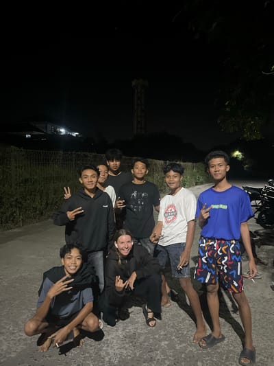In Labuhan Lombok, Indonesia, a group of eight young people, seven males and one female, are gathered outdoors at night. They are standing and crouching on a paved surface. The individuals are dressed casually, with the males wearing t-shirts and shorts, and the female in a dark hoodie and pants. Two of the males are making peace signs with their fingers. One male in the foreground is crouching and holding a cigarette. In the background, there is a chain-link fence, foliage, a building with lights on, and a tall, illuminated structure that appears to be a water tower. A few motorcycles are parked to the right of the group. The lighting suggests artificial illumination from nearby sources.