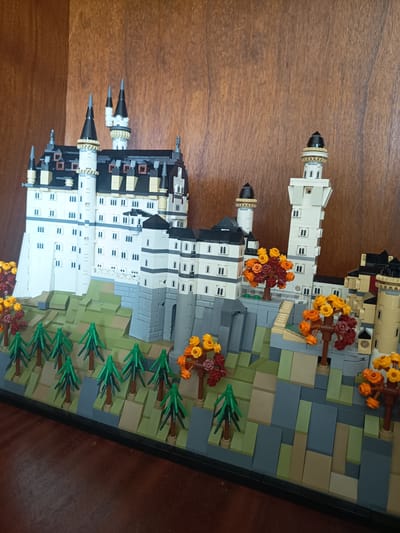 The image shows a detailed Lego model of a castle, likely Neuschwanstein Castle, set against a wooden background. The castle is predominantly white with black and grey accents, featuring multiple towers, spires, and intricate architectural details. The foreground of the model depicts a landscaped hillside with various shades of green, tan, and grey Lego bricks, representing terrain. Small green Lego trees, resembling pine trees, are planted on the hillside. Several Lego trees with orange and red foliage are strategically placed around the castle, suggesting an autumn theme. The Lego creation is displayed on a black base, which is placed on a wooden surface. The location in the city of Millstreet, Ireland is not evident from the image.
