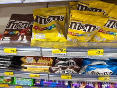 In the city of Wichit, Thailand, a brightly lit supermarket aisle showcases a variety of chocolate confectionery items displayed on two tiered shelves. The upper shelf is predominantly occupied by large bags of M&M's chocolates, including a dark brown bag labeled "milk chocolate" and several yellow bags featuring "peanut" and "m&m's" branding. Small M&M character illustrations are visible on some of the packaging. To the right, a yellow price tag indicating "79.00" and another labeled "12.00" are affixed to the shelf edge.

The lower shelf presents a selection of chocolate bars and wafer products. Prominently featured are several packages of Hershey's chocolate, including "Hershey's Kisses" and "Hershey's Chocolate Balls". A yellow price tag displaying "24.00" is visible on the left, while a tag with "38.00" is seen on the right. Beneath these, more confectionery items in various colored wrappers, including purple and green, are partially visible. The overall environment suggests a well-stocked retail setting.