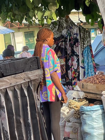 A woman with brown braids wearing a colorful patterned shirt lies atop a corrugated metal structure. Above her, colorful fabrics hang, creating a makeshift awning. A bag of nuts, sticks, and sacks sit nearby. The background reveals glimpses of buildings, trees, and other people. The scene appears to be an outdoor market or street setting.