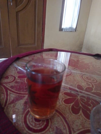 The image shows a glass of dark red liquid, likely tea, on a patterned table. A red cloth is draped over the edge of the table. In the background, a wooden door and a window with white curtains are visible. The scene appears to be indoors, possibly in a home in Sukamanah, Indonesia. The lighting is dim, suggesting it might be evening or the room is not well-lit. There are no people or animals visible in the image. The overall impression is a quiet, domestic setting.