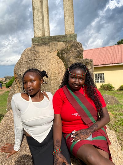 Two young women are posed on large rocks outdoors, with a building and cloudy sky in the background. The woman on the left is lying on her back, wearing a red shirt with green and dark red stripes across her chest and a matching skirt. She has black curly hair and is wearing a beaded headband in green and red. Intricate henna designs adorn her right hand and forearm. She is smiling and looking towards the camera.
The woman on the right is lying on her side, wearing a white long-sleeved top with a textured pattern. She has her hair braided into two buns and is looking towards the camera with a slight smile.
In the background, a building with a yellow facade and a red, corrugated roof is visible. A window with white bars is also present on the building. A concrete structure, possibly a pillar, is on the left side of the frame. The ground is covered in grass and rocks. The sky is overcast with dark clouds. The image appears to be taken during the daytime. There is text on the red shirt of the woman on the left which reads "NURSES OF H".