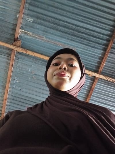 A low-angle, medium shot captures a young woman wearing a dark brown hijab and sweater, looking up towards the camera. The background consists of a corrugated metal roof supported by wooden beams, suggesting an outdoor or semi-outdoor setting. The woman's face is partially illuminated, revealing light makeup on her eyes and lips. The environment appears rustic and utilitarian. The location is specified as being in the city of Watampone, Indonesia, a region known for its cultural heritage and coastal landscapes.