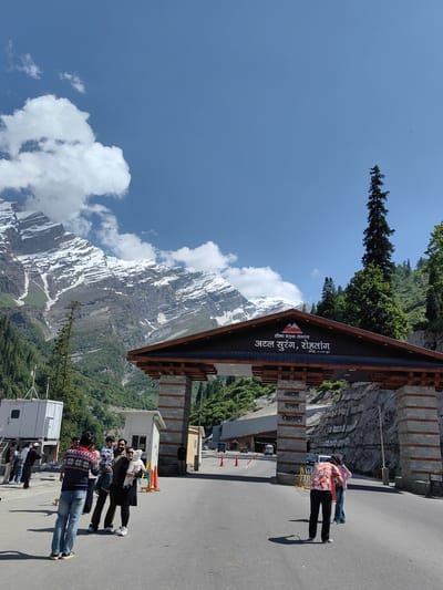 The image captures the entrance to the Atal Tunnel in Burwa, India, under a clear blue sky with scattered clouds. Majestic snow-capped mountains form a dramatic backdrop, with lush green trees covering the lower slopes. The tunnel entrance is an impressive architectural structure featuring a wooden roof supported by stone pillars. Text in Hindi is visible on the structure, indicating it is the "Atal Tunnel, Rohtang."
In the foreground, several people are present. Some are gathered near the entrance, possibly tourists. One individual in a colorful sweater and jeans is facing away from the camera, seemingly taking a photograph. Further into the frame, a family group stands near a smaller building. A man holds a child, and a woman holds a baby. Other individuals are seen walking or standing around the entrance area, with vehicles and traffic cones visible on the road leading to the tunnel. The scene appears to be a popular tourist spot, with visitors enjoying the mountain scenery and the engineering marvel of the tunnel.