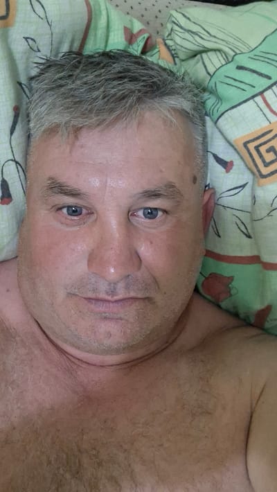 A close-up selfie of a middle-aged Caucasian male with graying hair, blue eyes, and stubble is taken from a high angle. The subject is lying down, with a pillow with a green and white pattern beneath his head. The man is bare-chested. The image appears to be taken indoors, likely in a bedroom in Voinova, Moldova.