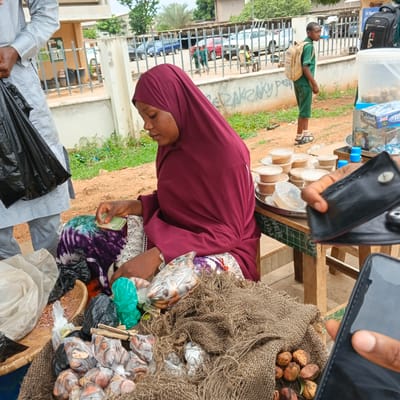 The image depicts a street scene in Shaba, Nigeria, where a woman wearing a maroon hijab and a patterned skirt is seated behind a display of goods. She is holding a small stack of Nigerian Naira currency in her right hand, appearing to be in the process of a transaction. In front of her are various items for sale, including small plastic containers of what appear to be spices or powders, and bags of nuts or seeds. A woven mat and burlap sacks are also visible, forming part of her stall. To the left, a person in a white and grey traditional outfit stands holding a black plastic bag. In the background, a young boy in a green school uniform with a backpack stands looking to his right, while other people and vehicles are visible behind a metal fence. The environment appears to be an outdoor market with a paved area and some greenery.