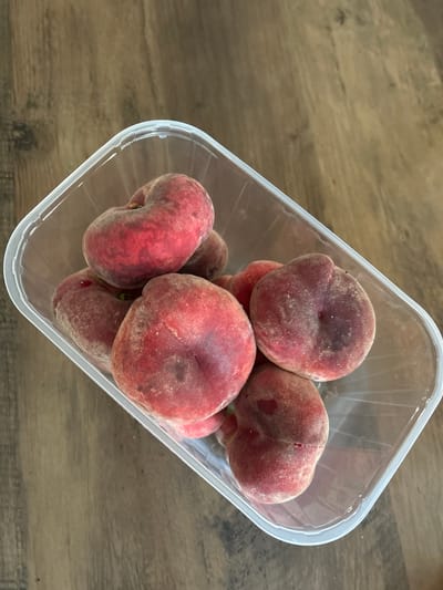 A clear plastic container filled with several flat peaches sits on a wooden surface. The peaches are a mix of red and pink hues, with a fuzzy texture. The container has a clear lid that is partially ajar. The wooden surface appears to be a table or countertop with a visible grain pattern. The lighting suggests an indoor environment, likely a kitchen or pantry. The scene is static, with no apparent action or people present. The overall impression is one of fresh produce, possibly prepared for consumption or storage. The image was taken from a slightly elevated angle, looking down into the container.
