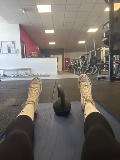 The image depicts a first-person perspective from an individual resting on a blue exercise mat within a gym located in Neuville-aux-Bois, France. The individual's legs, clad in black leggings and white high-top Converse sneakers with white socks, extend into the foreground. A black kettlebell with a teal base ring rests between their feet on the mat. The floor consists of dark rubber gym mats.
In the midground, a white partition separates the immediate workout area from a reception-like space. Behind this partition, a vibrant pink wall is visible, with a dark counter and objects. To the far left, a white wall features a bulletin board, and below it, a light wooden bench with multiple pairs of shoes arranged underneath. To the right, various pieces of gym equipment, including weight machines and racks, extend into the background.
In the background, a person is partially visible near cardio equipment. The ceiling is white with evenly spaced square fluorescent lights illuminating the entire facility.