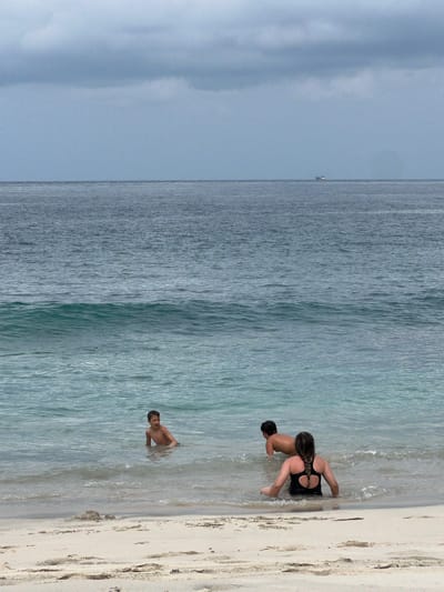 The image depicts a beach scene in Manggis, Indonesia, with three individuals in the shallow water. In the foreground, a woman in a black swimsuit with her hair in a braid is seated in the water facing away from the camera. To her left and slightly further out, a young boy with dark hair is crouched in the water, looking towards the left. Behind and to the left of the woman, another young boy with dark hair is on his stomach in the water, facing the same direction as the woman. The water is clear and calm with small waves. The beach in the foreground is sandy with some debris. The ocean stretches to the horizon, meeting a cloudy, grey sky. A distant boat is visible on the water.