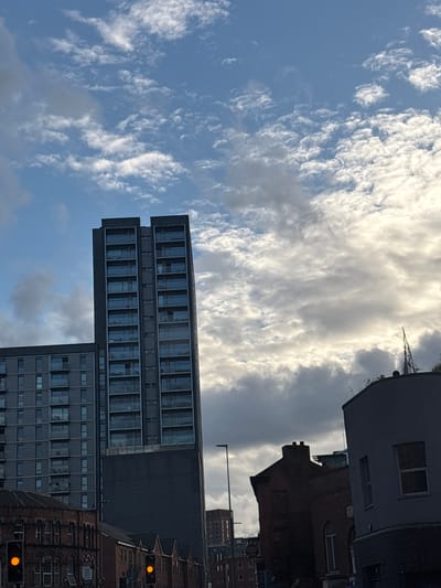 The image captures an urban street scene in Salford, United Kingdom. A tall, modern residential building with numerous glass-fronted balconies dominates the center of the frame. To its left, a shorter, similarly styled apartment building is visible. In the foreground, older brick buildings line the street, one featuring intricate architectural details and arched windows. Traffic lights are illuminated with amber signals. The sky above is a mix of blue and white clouds, with sunlight breaking through in some areas, suggesting either early morning or late afternoon. The overall impression is one of contemporary urban development alongside historic architecture.